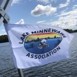 A white flag featuring the logo of the Lake Minnewawa Association, displaying a scenic graphic of a lake with boats and a colorful sunset.