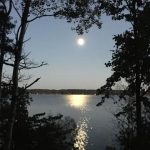 A serene lakeside scene at dusk with a full moon reflecting on the water, framed by trees.