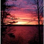 A vibrant sunset over a lake, with shades of pink, purple, and orange, framed by silhouettes of trees.