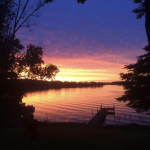 A serene sunset over a calm lake, with vibrant colors reflecting on the water and silhouettes of trees and a dock in the foreground.