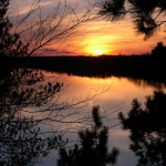 A serene sunset over a calm lake, framed by silhouetted trees and vibrant colors in the sky reflecting on the water.